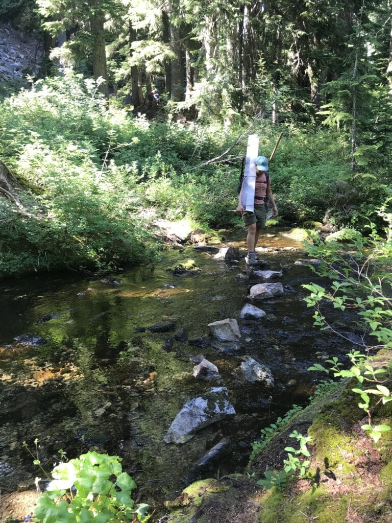 A person carrying a saw across a rocky creek.